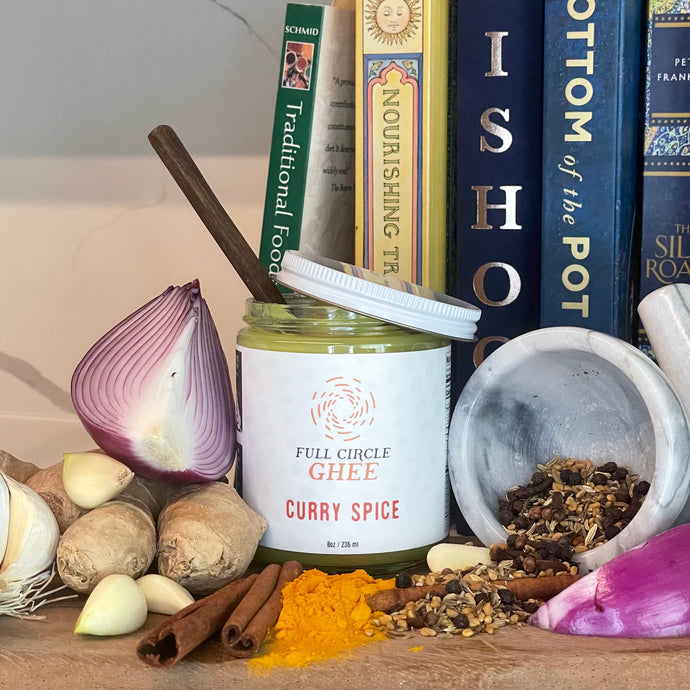 Curry Spice Full Circle Ghee arranged with cookbooks, spices, onions, garlic, and ginger.
